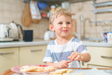 Little blond kid tries freshly baked shortbread, sitting at the kitchen table. Smiling face.