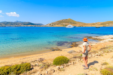 Young woman tourist standing on shore and looking at azure sea with sailing boats on coast of Paros island, Greece - obrazy, fototapety, plakaty