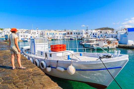 Young woman tourist standing on pier in front of a fishing boat in Naoussa port, Paros island, Greece