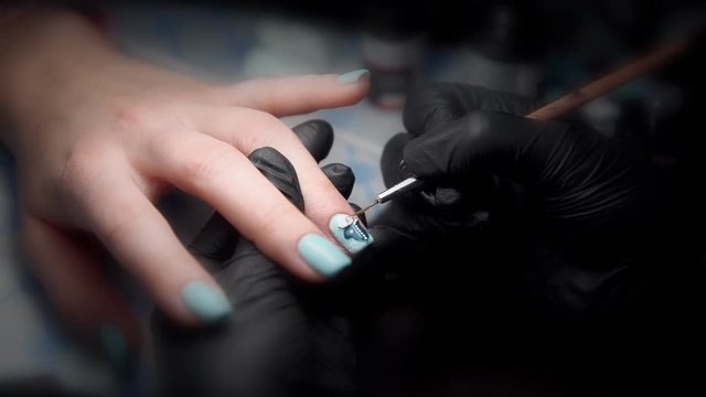 Manicurist in black gloves gets a top coating on the nail with drawing of a penguin shot close-up under bright light.