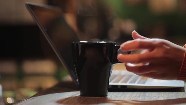 Panning footage of a person typing on a computer and grabbing a steamy coffee mug.