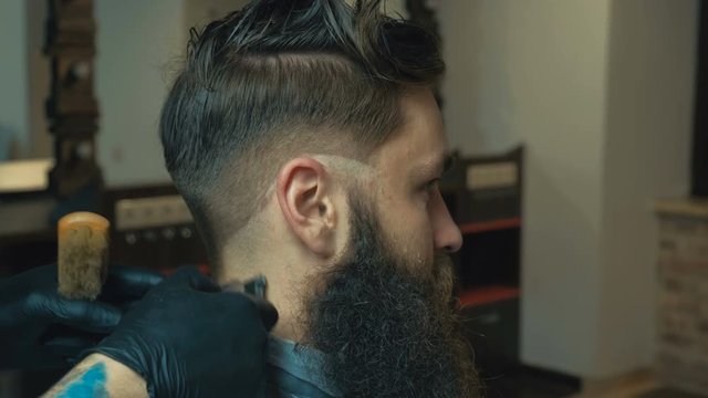Unparalleled barber with a beard and a tattoo is cutting the hair of his client in the barbershop. He is using a cutting comb and a hair clipper.