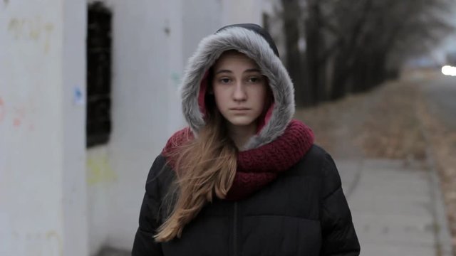 Young girl sadness and loneliness. Teenager wearing fur hooded jacket standing alone near road at old bus stop in cold evening, steadicam portrait shot.