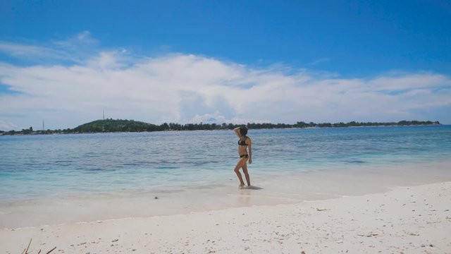 A tanned slim woman in black bathing suit walking on a sunny day on the edge of the ocean and the sand on the sandy islands of background