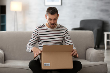 Handsome young man opening box with parcel while sitting on sofa at home