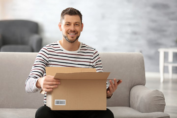 Handsome young man opening box with parcel while sitting on sofa at home