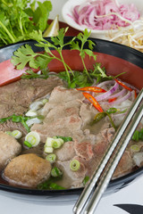 A bowl of traditional vietnamese beef and chicken pho street food, surrounded by fresh herbs. Pho is a vietnamese noodle soup consisting of meat, herbs, chilli, coriander and dumplings.