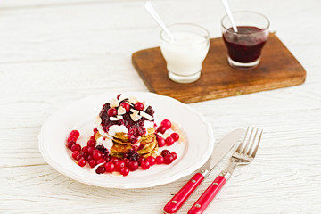 Stack of vegan gluten free pancakes with yogurt, blueberrty jam and canberries on a white plate with red fork and knife. Bright healthy breakfast. White wooden table