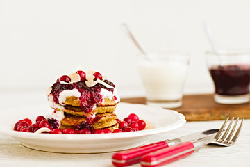 Stack of vegan gluten free pancakes with yogurt, blueberrty jam and canberries on a white plate with red fork and knife. Bright healthy breakfast. White wooden table