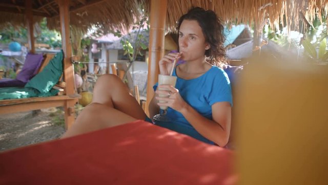 Young beautiful woman with black hair, dressed in a blue dress sitting on a sandy beach on a chair under a straw umbrella and thoughtfully drinking juce, looking far away