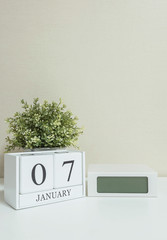White wooden calendar with black 7 january word with clock and plant on white wood desk and cream wallpaper textured background , selective focus at the calendar
