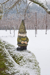 Gnome from backside looking into woods standing on snowy branch/Gnome with back to camera standing on snowy mossy tree branch looking out into snow covered woods