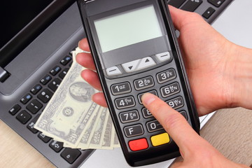 Hand of woman using payment terminal, currencies dollar on laptop in background