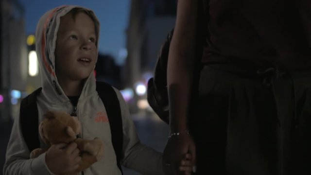 Slow motion steadicam shot of a boy with monkey toy holding mothers hand as they walking in the street at night