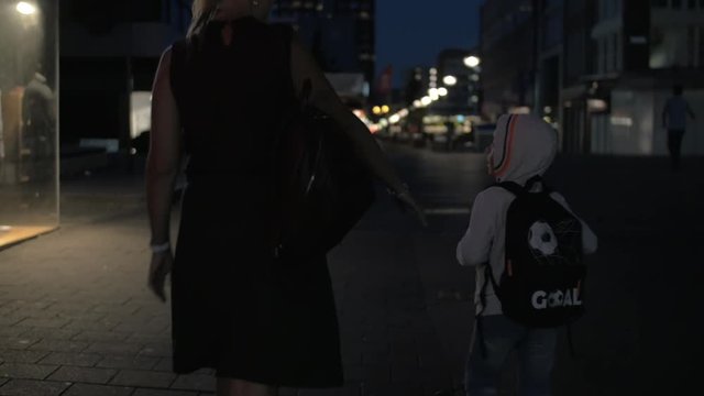 Slow motion view of woman with son going on the night empty pedestrian street, she is in black dress, he is with rucksack, Rotterdam, Netherlands