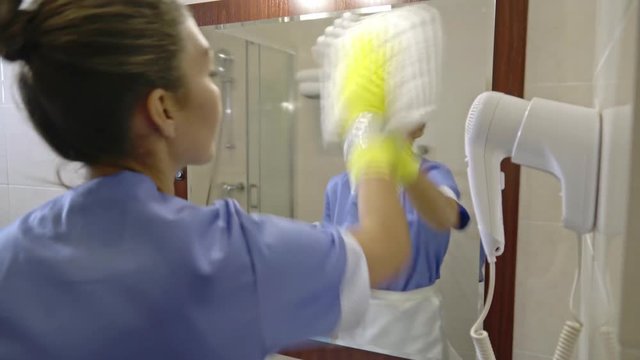 Concentrated Latin-American housekeeper spraying glass cleaner on mirror in hotel bathroom and wiping it with cotton rag