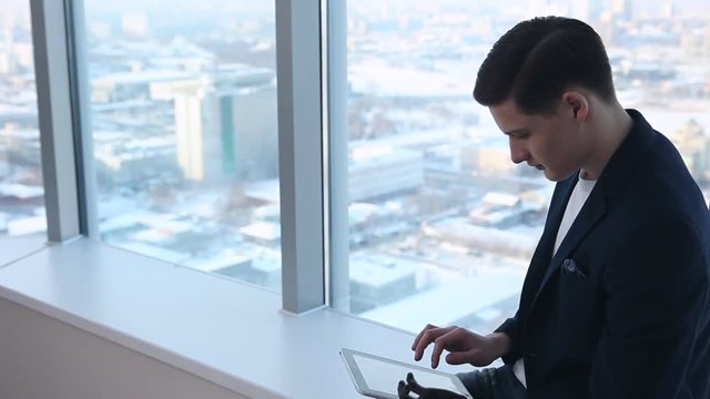 During a Break with tablet in the hands/Young student, manager, designer, architect sits on a background of a large window with a tablet in his hand