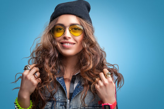 Closeup portrait of beautiful trendy hipster girl with curly hair and hands up smiling wearing red checkered shirt,denim vest and black beanie hat on blue background.Youth style,fashion.