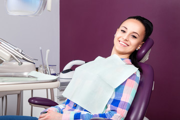 Young woman patient in checkered shirt with perfect straight white teeth waiting for dentist in dental chair and smiling relaxed, ready for a check-up. Beautiful woman smile