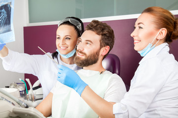 Obraz premium Smiling female doctor and her assistant showing teeth x-ray to male patient in dental hospital. Handsome male patient sitting in chair smilling with straight white teeth and looking at dental x-ray.