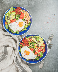 Healthy breakfast bowls with fried egg, chickpea sprouts, seeds, vegetables and greens over grey concrete background, top view, copy space. Clean eating, healthy lifestyle, vegetarian food concept
