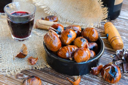 Delicious roasted chestnuts on ceramic bowl and glass of red wine on old wooden table, bottle of wine, fir cone, cork and vintage corkscrew in the background.