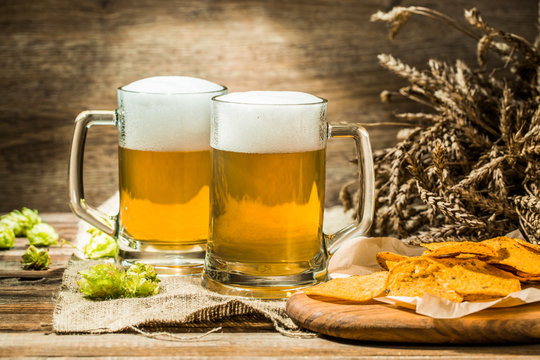 Two beer mugs with hops and chips on boards on linen cloth