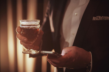 Young ginger bearded man holding a vintage glass with red wine against the light. black on background. guy looking in the phone