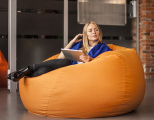 Armchair traditional bean bag for fun and relaxation  orange color. Young woman  using tablet screen while sitting on big cushioned frameless chair