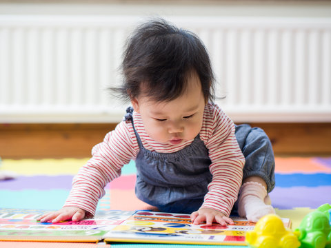 Adorable baby girl sitting on play mat  and reading book at home