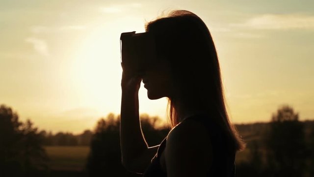 Woman uses a virtual reality glasses in the forest. Sunset light, sun lens flares, golden hour. Relax, nature and technology concept
