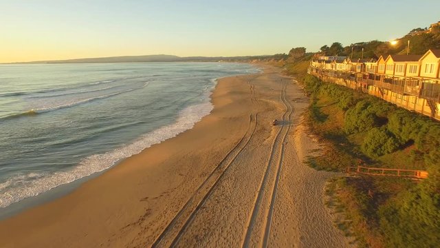 Aerial flying shot down a beach in California with beach houses during sunset. Golden sand, blue water and clear skies. Vehicle tracks in the sand. 