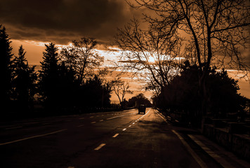 View of asphalt road  with car among dark silhouette of naked trees and  cloudy sunrise painted sepia