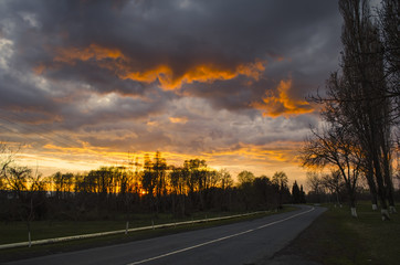 Obraz premium Landscapes of asphalt road in countryside among the silhouette of trees painted in sunset colors and covered dramatic sky with bright clouds.