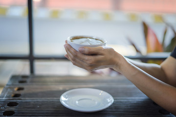 Female hands holding a cup of coffee with foam over wooden table, side view with soft focus blur background