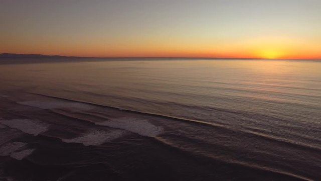 Slow panning aerial shot at the beach in California during sunset. Sharp horizon line, golden/orange/red light of the sun. Deep blue ocean color and clear skies. 
