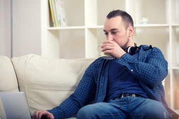 Handsome man sitting on sofa with laptop drinking tea