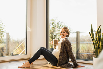 Smiling pregnant woman sitting on floor enjoying a drink in mug