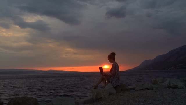 Elegant girl sitting on the shore of the Adriatic Sea at sunset and typing on leptope. Woman working on the beach as freelancer. The life of a girl blogger of the sea