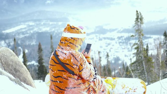 young girl on a snowboard in a suit looks tigers photo on the phone sitting on a snow-covered mountain. 4k