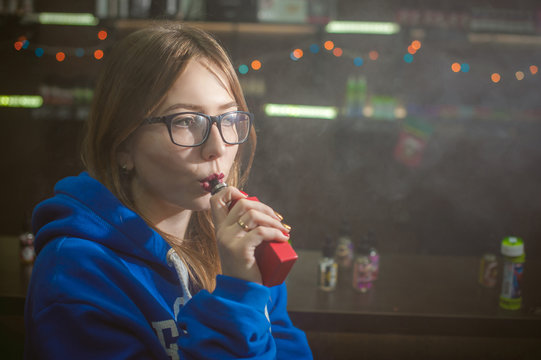 beautiful young woman with glasses smoking electronic cigarette. girl in a blue sweater brings to his lips red box mod, sitting indoors vape shop cafe