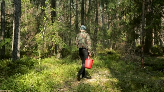 young girl in the khaki jacket and a bandana on your head goes through a dense forest and looking for mushrooms with a red bucket in his hand.