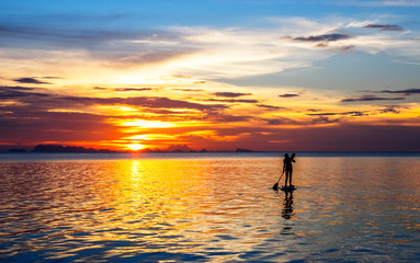 Silhouette of a young woman paddleboarding at sunset with cloudy landscape on the background