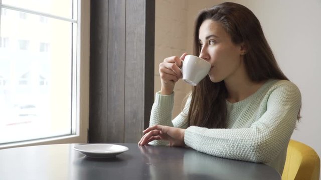 Beautiful girl sitting at a table and drinking tea. Dreamed eyes looking out the window.