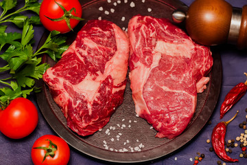 Raw meat, beef Ribeye and tomatoes, parsley, spices on a round cutting board. Dark background, top view