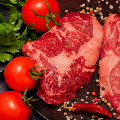 Fresh raw marbled steaks chops on black background with tomatoes, parsley and spices. Square