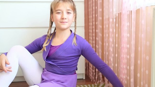 Beautiful little ballerina in lilac dress for dancing sitting on the floor looking at camera and smiling