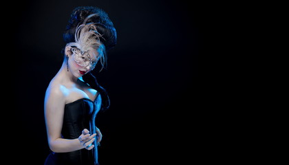 brunette woman with high hair, a mask with feathers and corset in old style on a black background