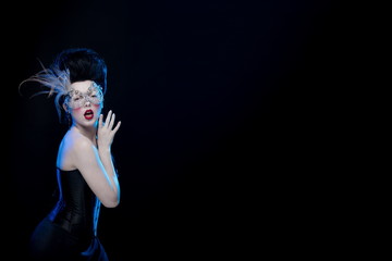 brunette woman with high hair, a mask with feathers and corset in old style on a black background