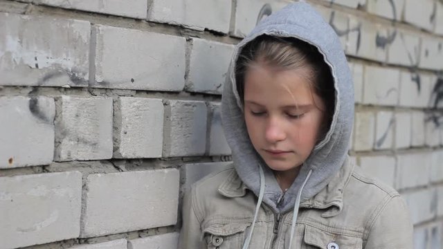 Sad young girl leaning head against brick wall looking down. Portrait of depressed kid wearing grey hooded jacket standing alone in city.